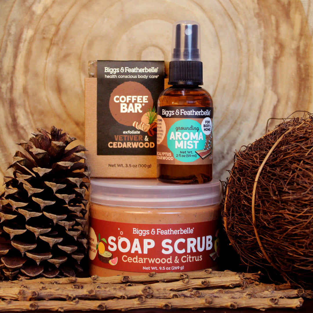 Three natural beauty products including a soap scrub, coffee bar, and aroma mist on a wooden surface with a pine cone and coir ball.