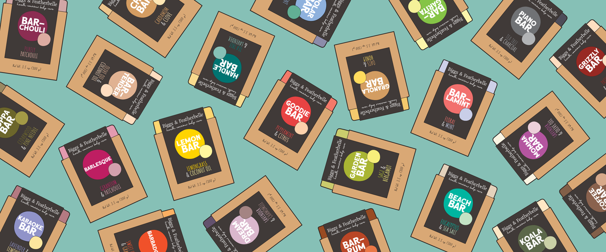 A repeating pattern of illustrated packaged soap bars on a light aqua background. The packages are tan with dark brown labels and colorful flavor icons, including orange, yellow, green, pink, blue, and purple.