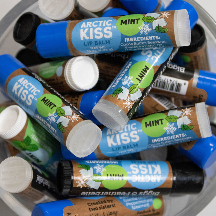 Several Arctic Kiss Lip Balm tubes in Mint flavor are piled together in a clear container. The tubes are blue with brown and green label graphics, white caps, and visible ingredients including cocoa butter, beeswax, and peppermint oil.