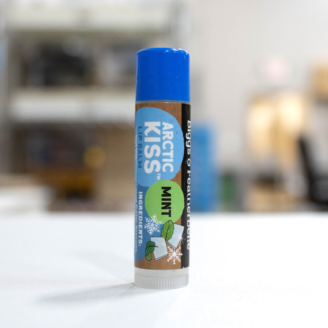 A single tube of B&F lip balm standing upright on a white surface with a blurred indoor background. The product is Arctic Kiss Mint, featuring a blue cap, brown label, and blue, green, and white winter-themed design.