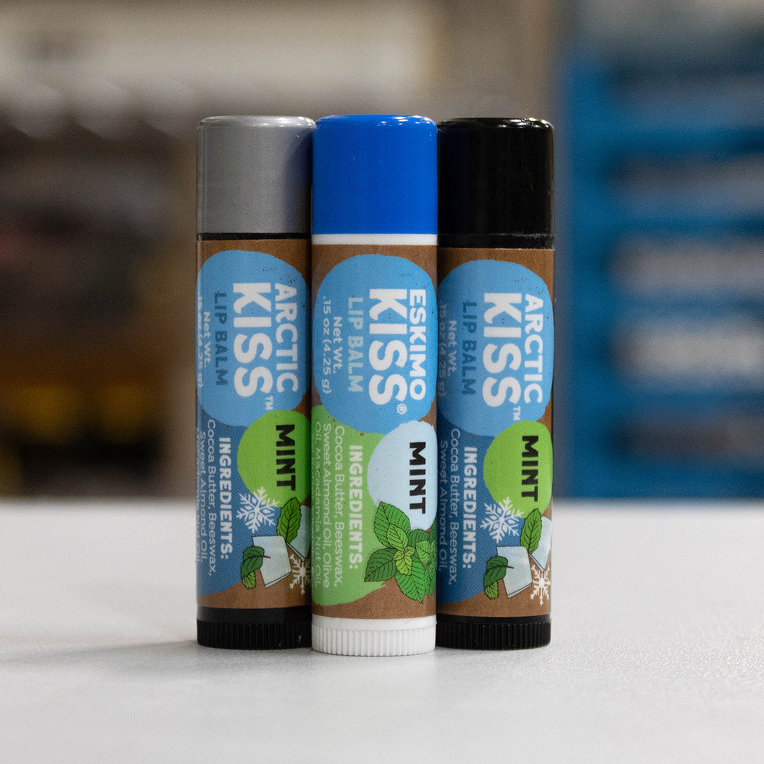 Three Arctic Kiss Lip Balm tubes standing upright on a white surface, with a blurred indoor background. The center tube is mint green and white with a blue cap, and the side tubes are brown with black and silver caps.