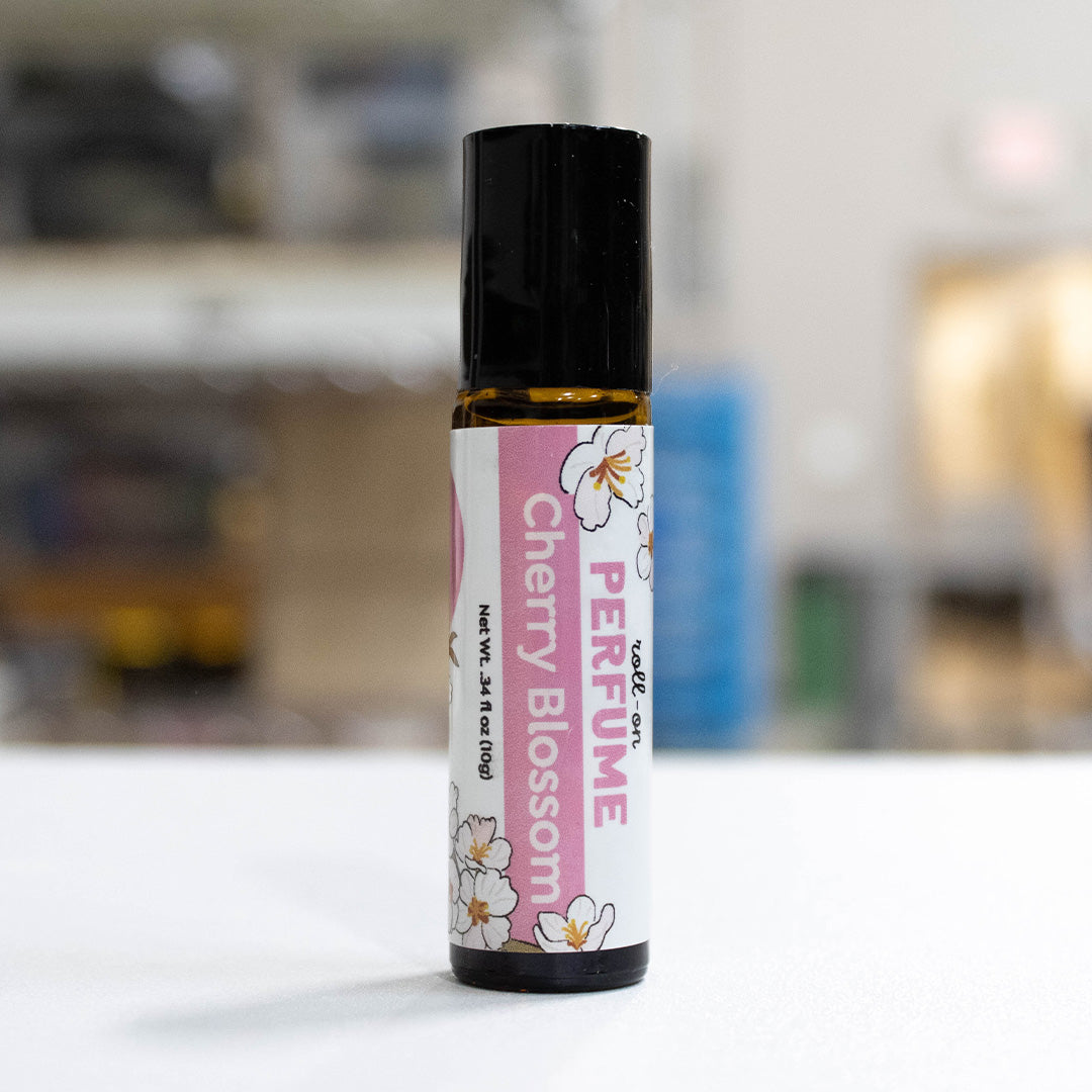A small amber glass perfume roller bottle with a black cap and a pink-and-white B&F label reading 'PERFUME Cherry Blossom' with cherry blossom illustrations, standing on a white surface with a blurred indoor background.