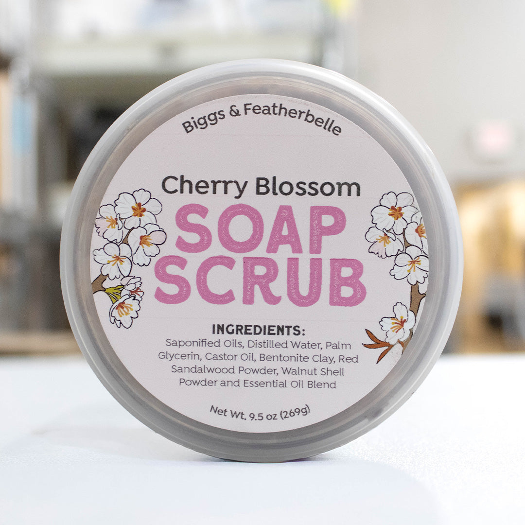 A round jar of Biggs & Featherbelle Cherry Blossom Soap Scrub, featuring a white label with pink lettering and cherry blossom illustrations, set against a softly blurred background.