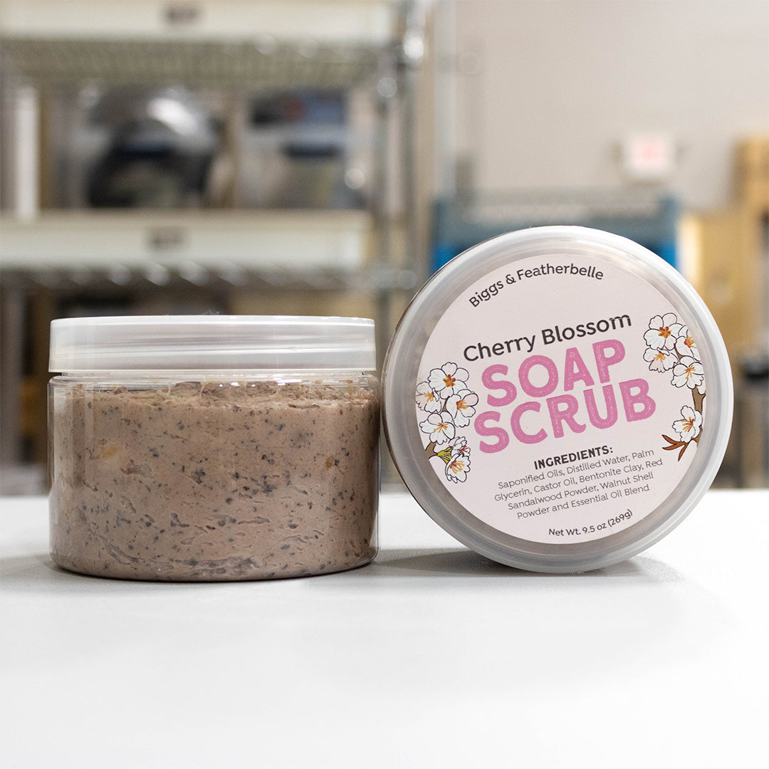 A jar of Biggs &amp; Featherbelle Cherry Blossom Soap Scrub shown next to its lid on a white surface, with a softly blurred industrial background.
