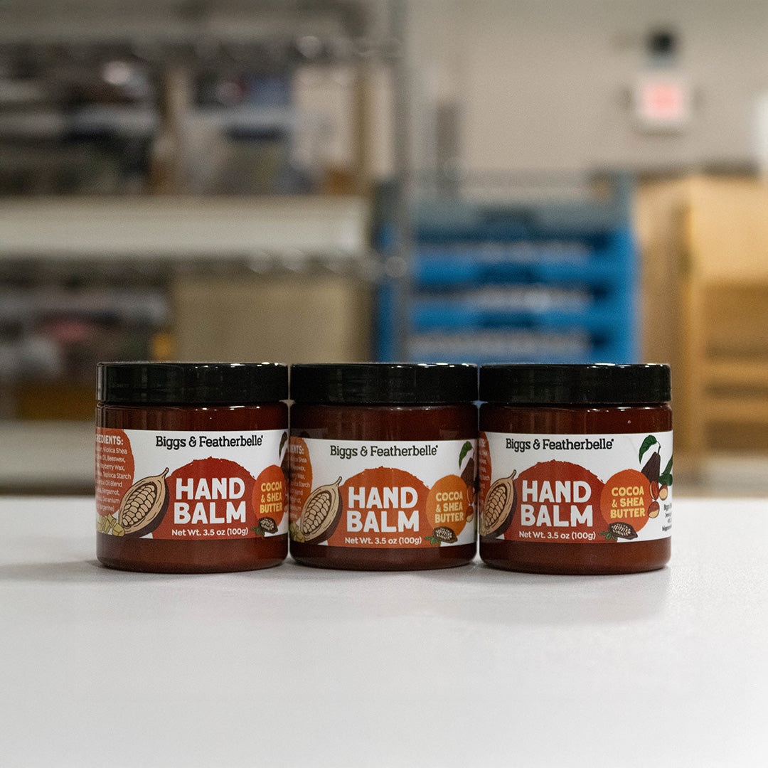 Three jars of Biggs &amp; Featherbelle Hand Balm, specifically Cocoa &amp; Shea Butter, lined up on a white surface with a blurred warehouse or storage room background.