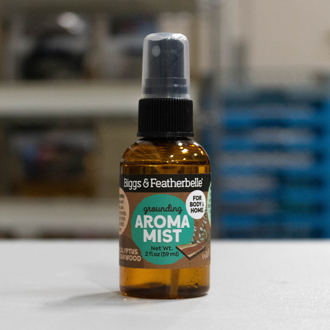 A 2 fl oz amber glass spray bottle of Biggs &amp; Featherbelle Grounding Aroma Mist, featuring a black spray cap and a label with green and white graphics, set against a blurred indoor background.