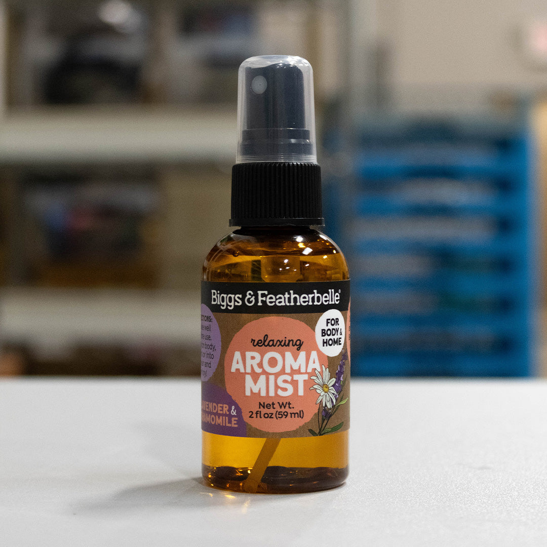 A bottle of Biggs &amp; Featherbelli Relaxing Aroma Mist in lavender and chamomile, featuring an amber glass bottle, black spray cap, and a purple and orange label, set against a blurred indoor background.