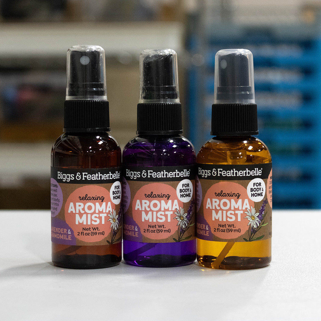 Three bottles of Biggs & Featherbell relaxing Aroma Mist lined up on a white surface. The left bottle is amber with a black spray top, the middle bottle is purple with a black spray top, and the right bottle is golden yellow with a black spray top.