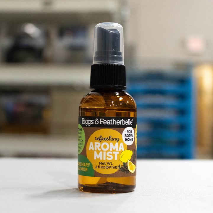 A bottle of Biggs &amp; Featherbelle Freshing Aroma Mist in German Lemon scent, featuring an amber glass bottle with a black spray cap and a green and yellow label, set against a blurred background.