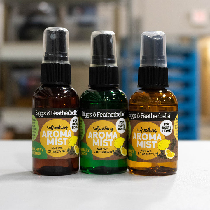 Three Biggs &amp; Featherbelle refreshing Aroma Mist spray bottles lined up on a white surface with a blurred indoor background. The bottles are amber brown, green, and clear with black spray caps, featuring yellow and green labels.