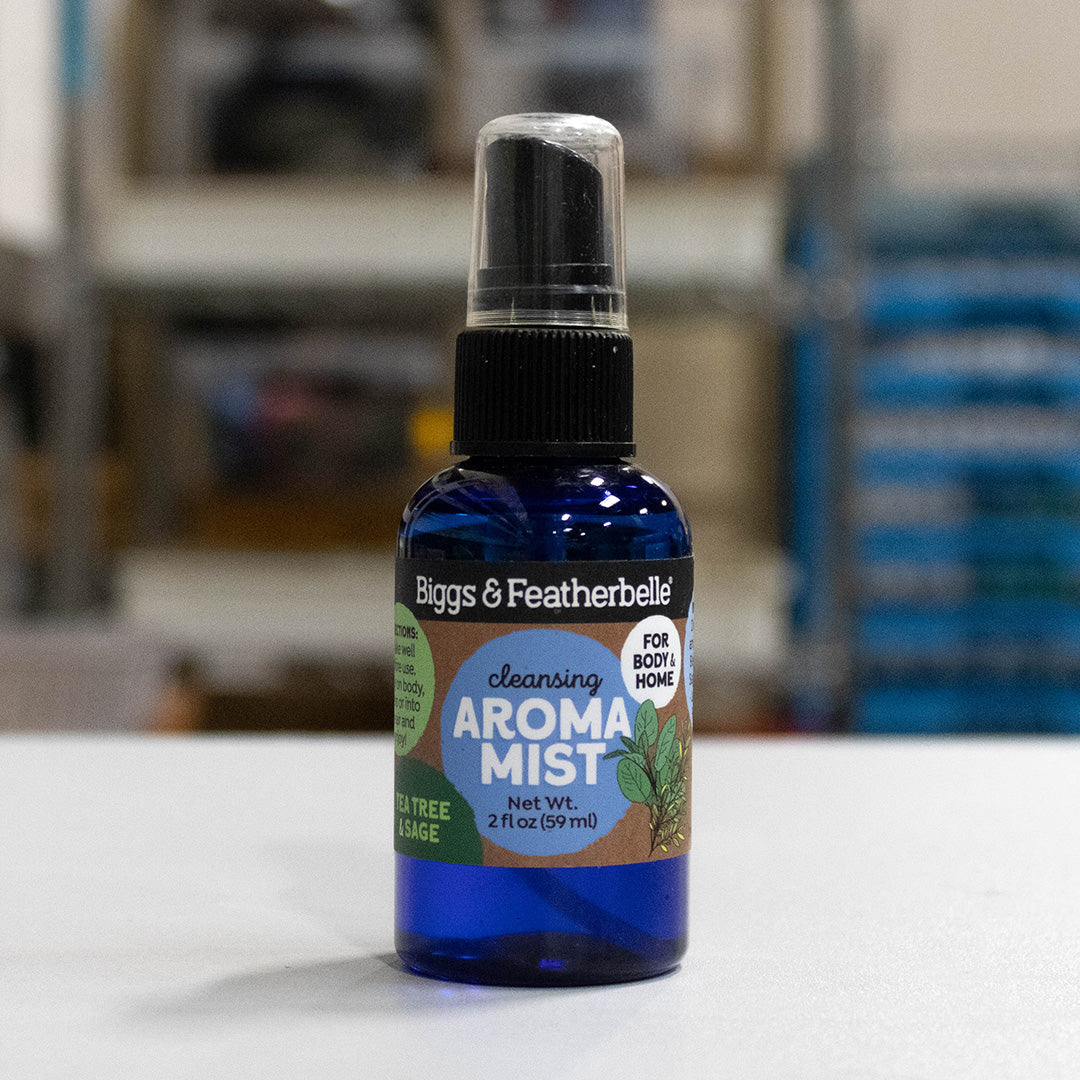 A 2 fl oz blue glass spray bottle of Biggs &amp; Featherbelli Cleansing Aroma Mist, featuring a black spray cap and a colorful label, set against a blurred indoor background.