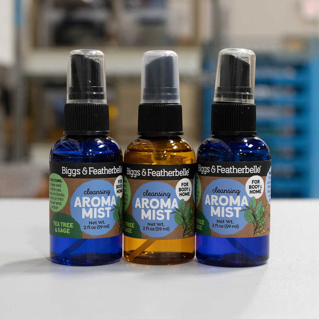 Three bottles of Biggs & Featherbelle Cleansing Aromat Mist lined up on a white surface with a blurred indoor background. The left bottle is blue with a green label, the middle bottle is amber with a green label, and the right bottle is cobalt blue with a light blue label.