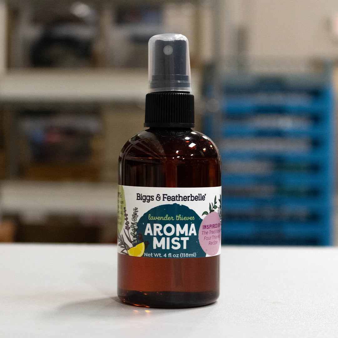 A 4 fl oz amber glass spray bottle of Biggs & Featherbelle Lavender Thieves Aroma Mist, standing on a white surface with a blurred indoor background.