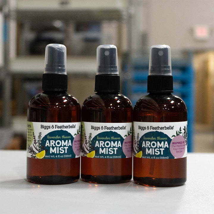Three amber glass spray bottles of Biggs &amp; Featherbelle Aroma Mist with black spray tops, lined up on a white surface with a blurred workshop or storage room background.