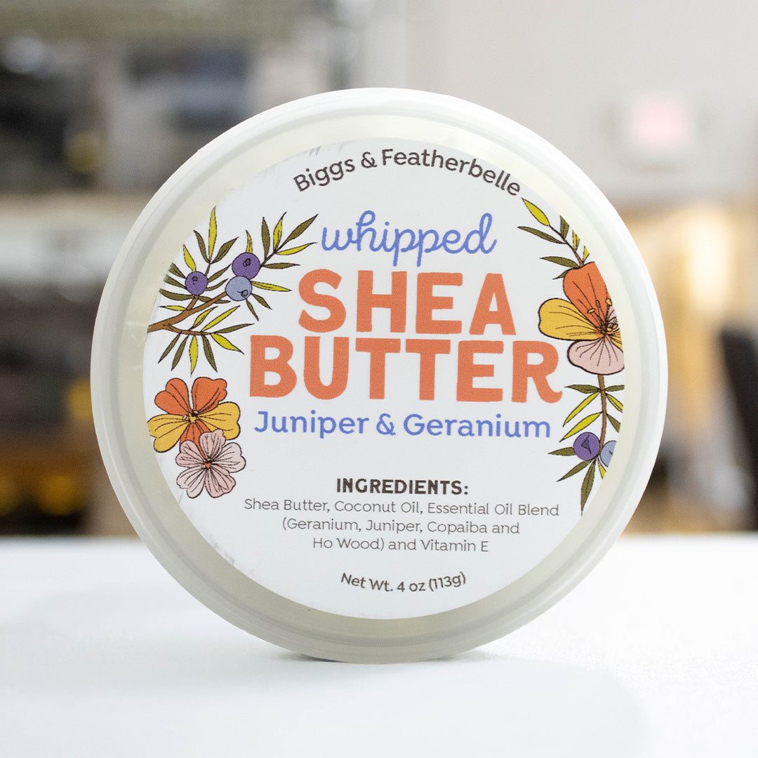 A round white tub of Biggs & Featherbelle whipped Shea Butter, Juniper & Geranium scent, shown from a top-down view on a white surface with a blurred background.