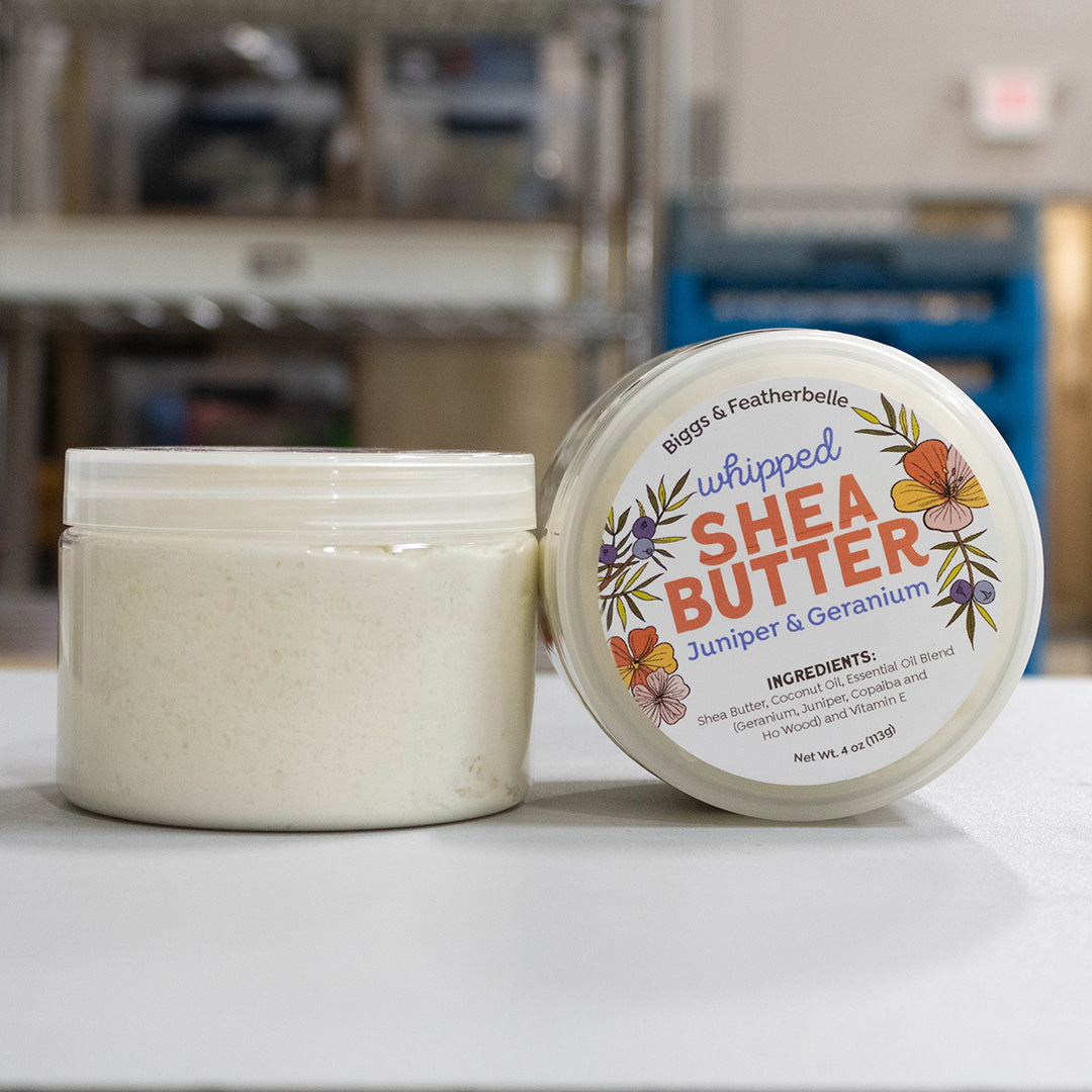 An open jar and matching lid of Biggs &amp; Featherbelle Whipped Shea Butter on a white surface, with a softly blurred indoor background.