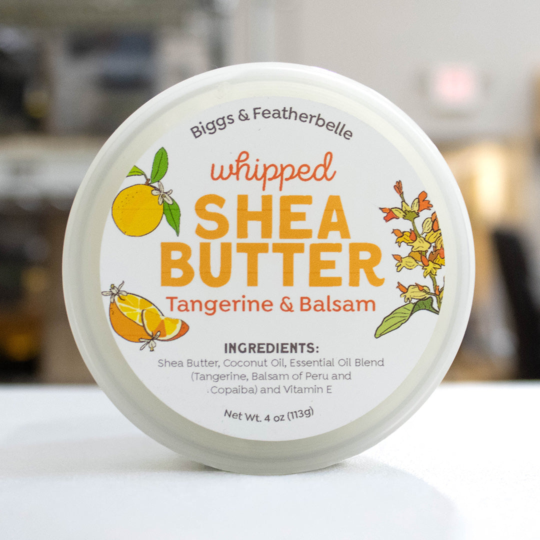 A round white plastic tub of Biggs & Featherbelle Whipped Shea Butter in Tangerine & Balsam, featuring a white lid with orange and yellow illustrations of tangerine and a balsam flower, set against a blurred background.