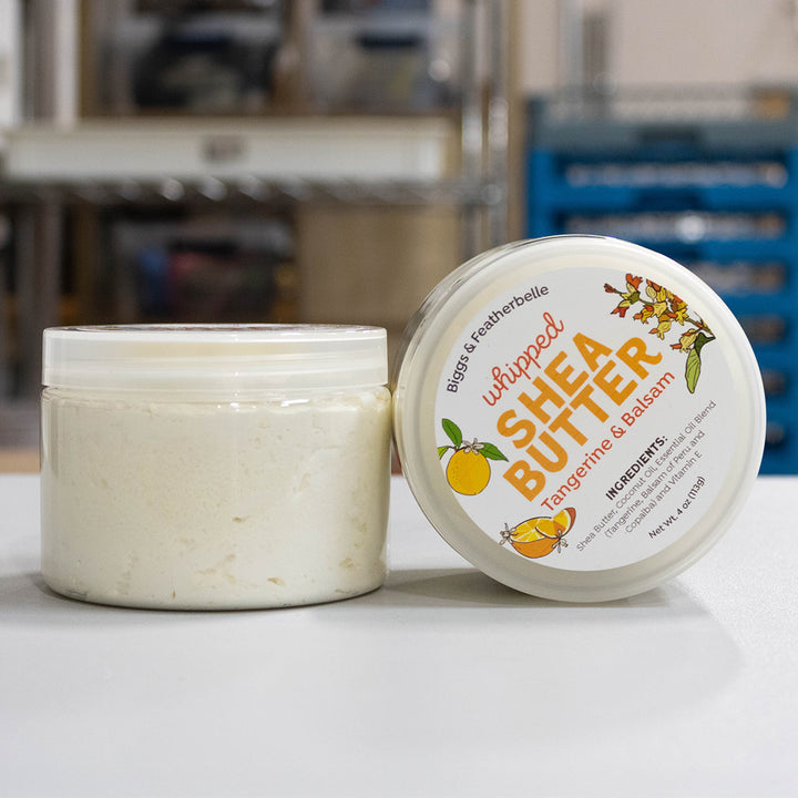 Two jars of whipped shea butter on a white surface, with one jar open showing the off-white cream inside and the other showing the lid label. The label reads &