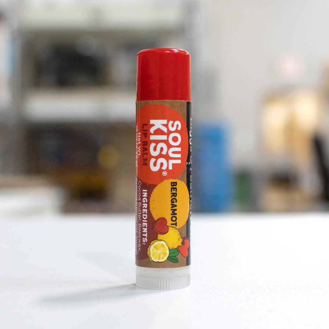 A single tube of B&amp;F Soul Kiss Bergamot Lip Balm standing upright on a white surface with a blurred indoor background.