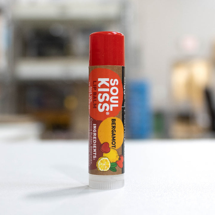 A single tube of B&amp;F Soul Kiss Bergamot Lip Balm standing upright on a white surface with a blurred indoor background.