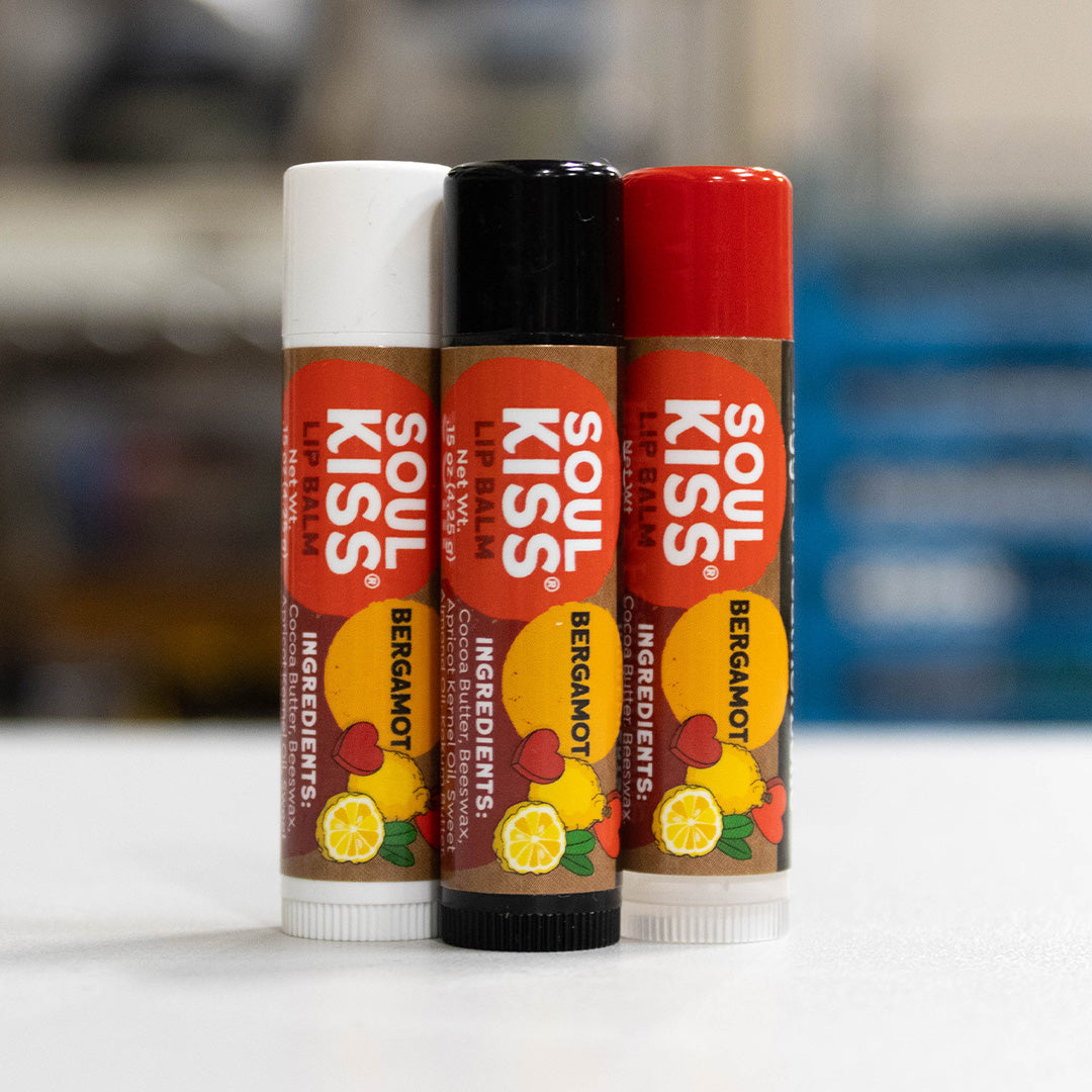 Three tubes of Soul Kiss Bergamot Lip Balm lined up on a white surface with a blurred background. The lip balms have white, black, and red caps, and the labels feature orange, yellow, and red graphics with illustrations of lemons, bergamot, and berries.