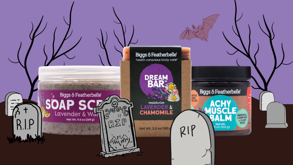 Three Biggs & Featherbelle body care products displayed together against a purple Halloween-themed background with tombstones, bare trees, and a bat. The products include a lavender and witch soap scrub jar, a lavender and chamomile dream bar box, and an achy muscle balm jar.