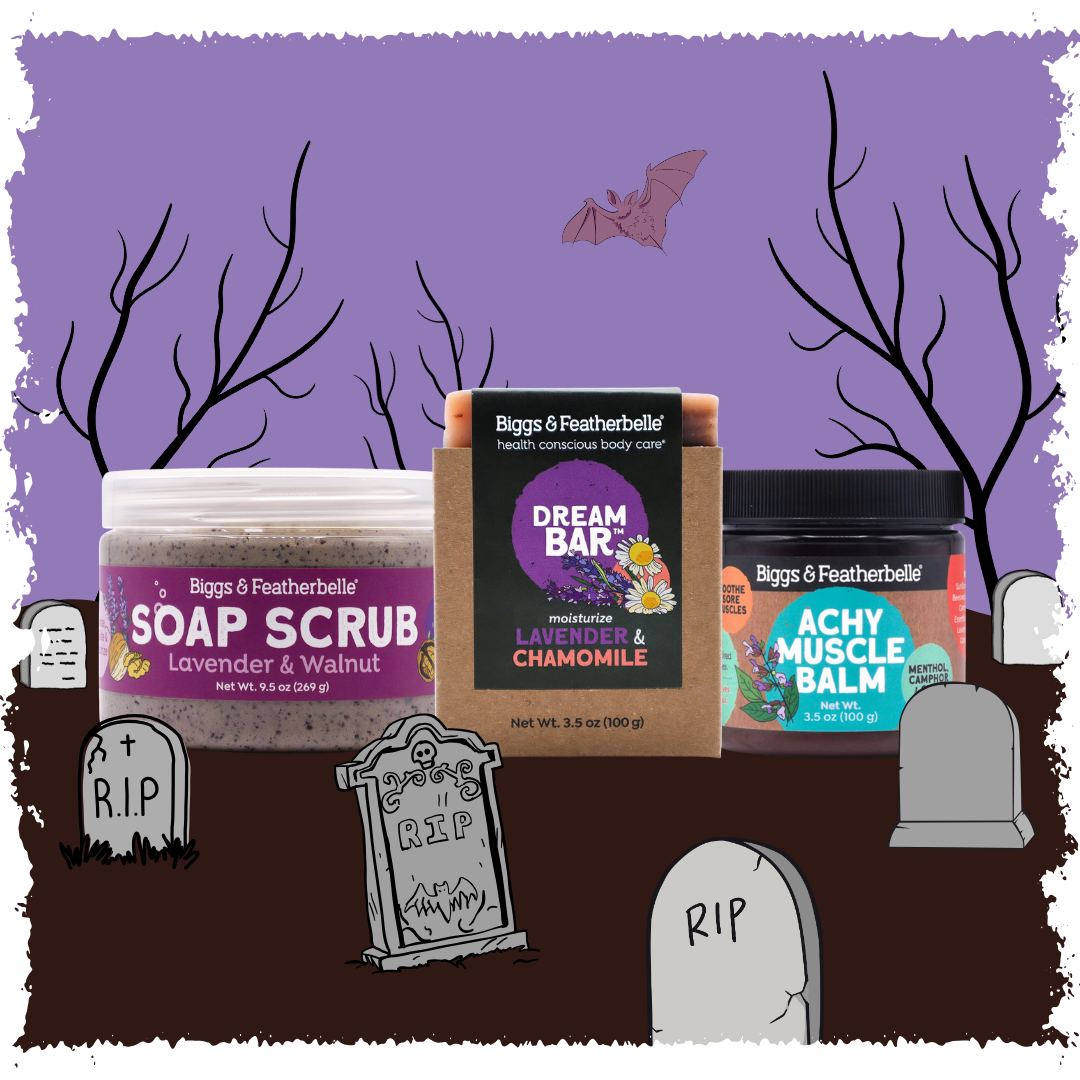 Three Biggs & Featherbell bath products arranged together against a Halloween-themed illustrated background with tombstones, bare trees, a bat, and a purple sky. The products are Soap Scrub, Dream Bar, and Achy Muscle Balm.