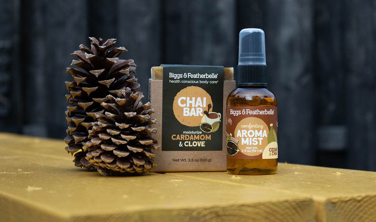 Biggs & Featherbelle Chai Bar aromatherapy set featuring a pinecone, a cardboard box of Cardamom & Clove Chai Bar soap, and a bottle of Aroma Mist on a wooden surface against a dark wood background.