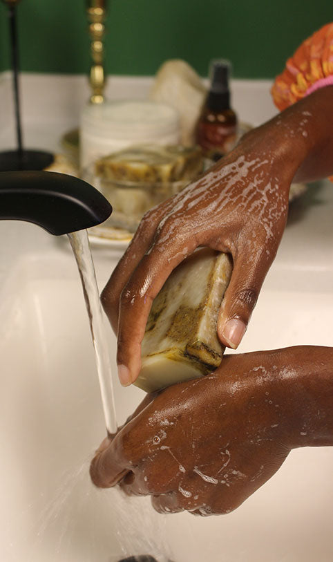 A close-up of a person's hands washing a bar of soap under running water in a white sink, with toiletries blurred in the background.